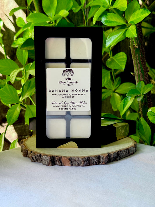 A black box with a clear plastic tray housing eight white wax melts featuring a white label with black writing on a wooden surface with green leaves in the background