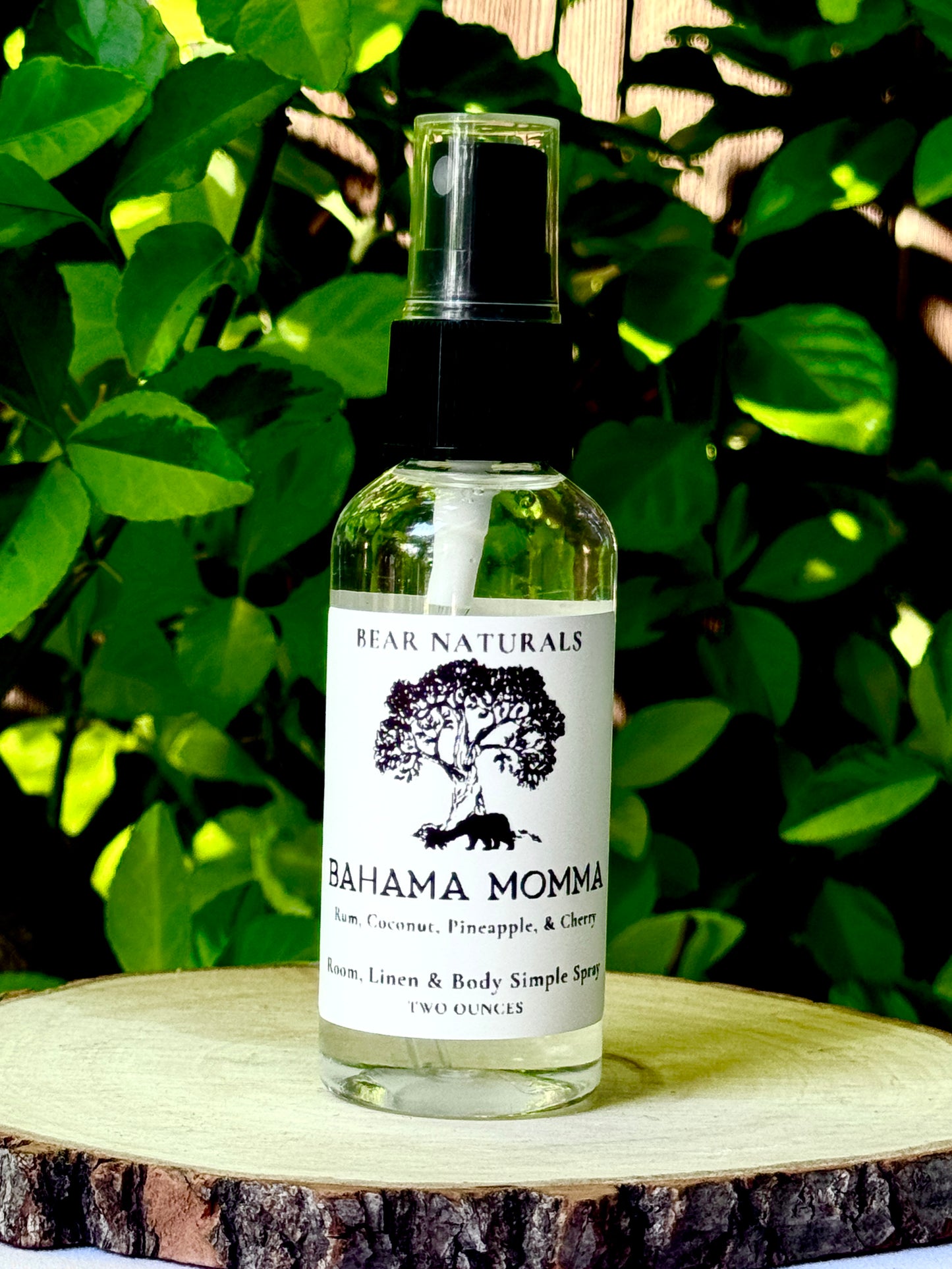 A clear bottle with a black fingertip style spray nozzle featuring a white label with black writing on a wooden surface with green leaves in the background.