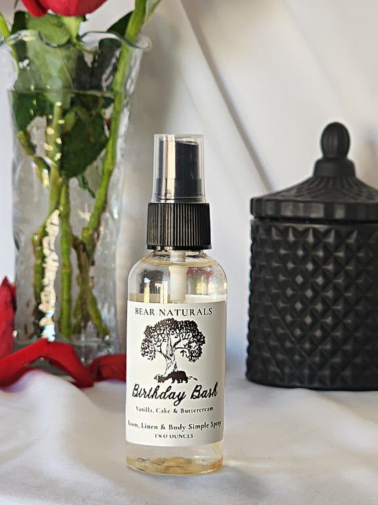 A clear bottle with a black fingertip style spray nozzle featuring a white label with black writing with a black candle jar and red roses in a vase in the background.