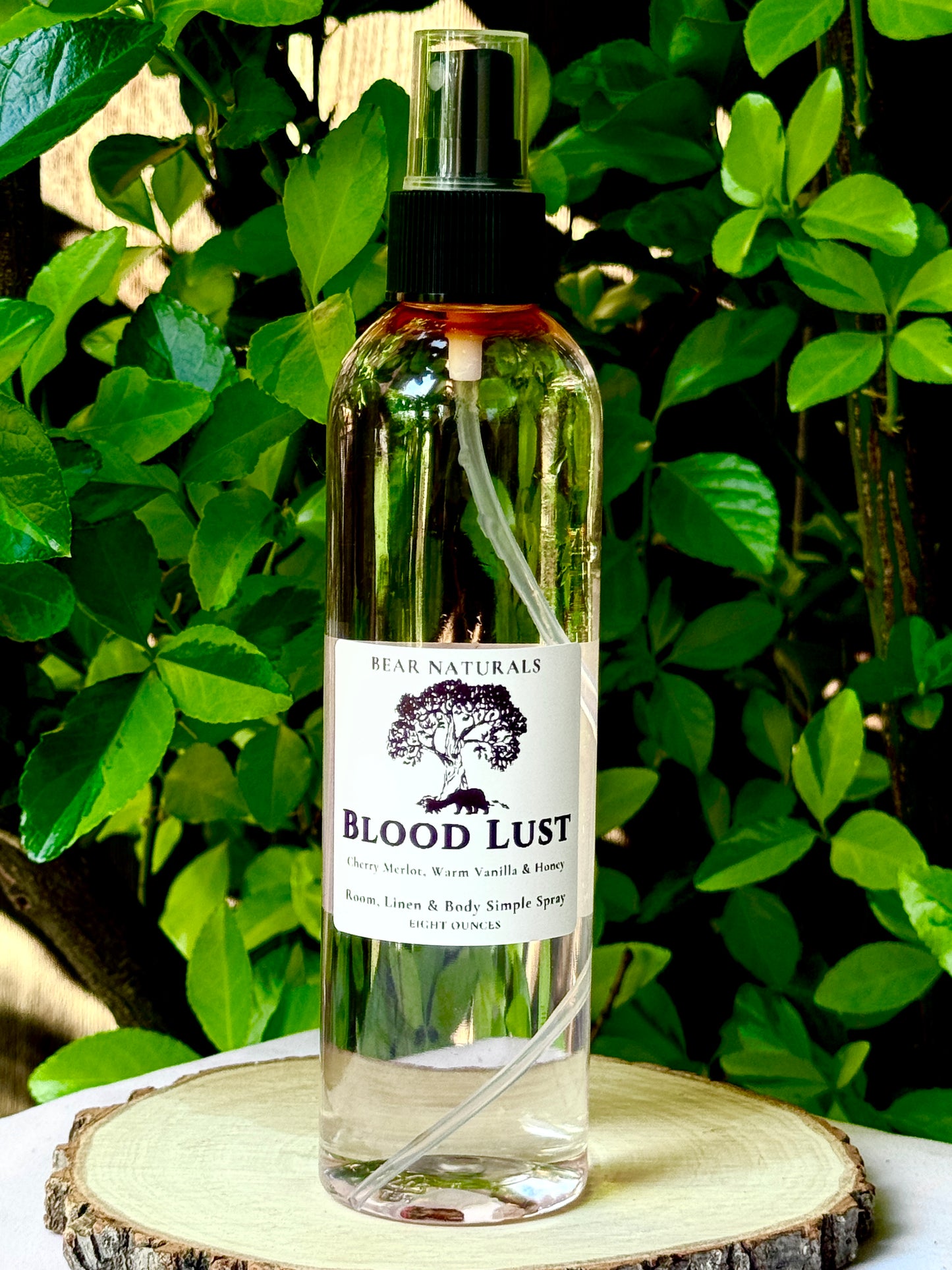 A clear bottle with a black fingertip style spray nozzle featuring a white label with black writing on a wooden surface with green leaves in the background.
