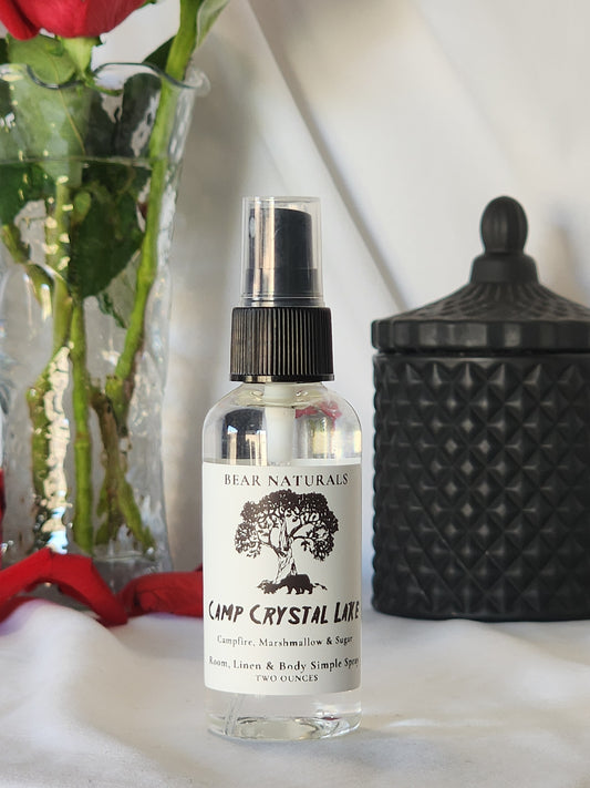 A clear bottle with a black fingertip style spray nozzle featuring a white label with black writing with a black candle jar and red roses in a vase in the background.