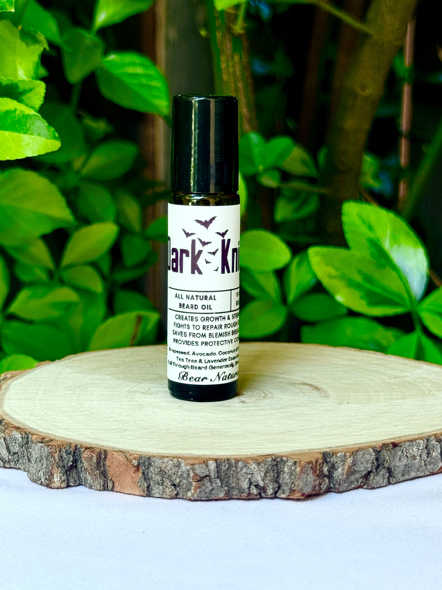 10ml amber colored roll on bottle with a black lid featuring a white label reading "Dark Knight All Natural Beard Oil" sitting on a wooden surface with green leaves in the background