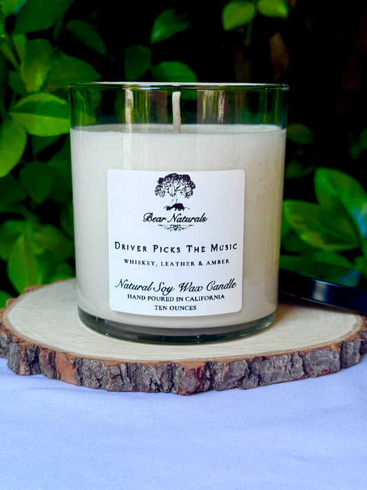 a ten ounce white candle in a clear jar featuring a white label with black writing on a wooden surface with green leaves in the back ground.