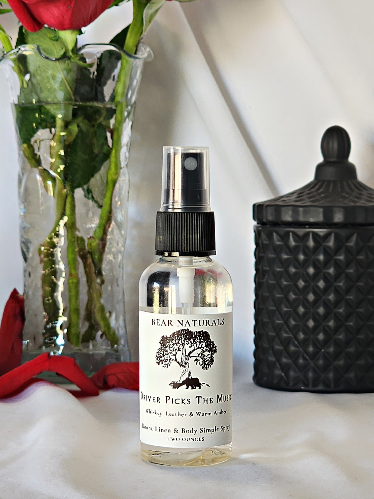 A clear bottle with a black fingertip style spray nozzle featuring a white label with black writing with a black candle jar and red roses in a vase in the background.