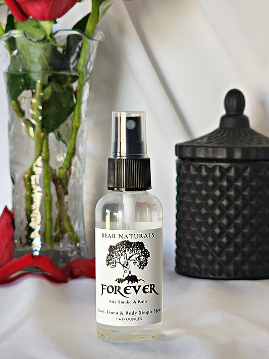 A clear bottle with a black fingertip style spray nozzle featuring a white label with black writing with a black candle jar and red roses in a vase in the background.