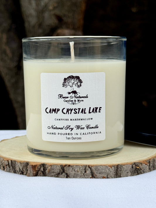 A ten ounce white scented candle in a clear glass jar with a black metal lid and white label with black writing sitting on a wooden surface. The candle is called "Camp Crystal Lake".