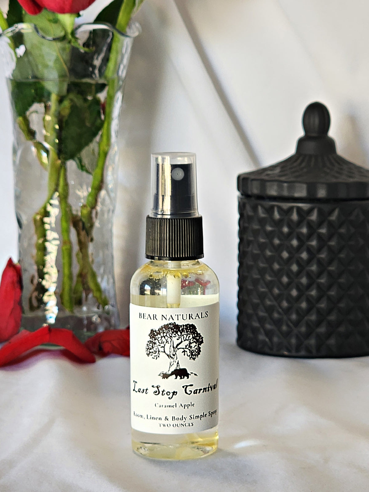 A clear bottle with a black fingertip style spray nozzle featuring a white label with black writing with a black candle jar and red roses in a vase in the background.
