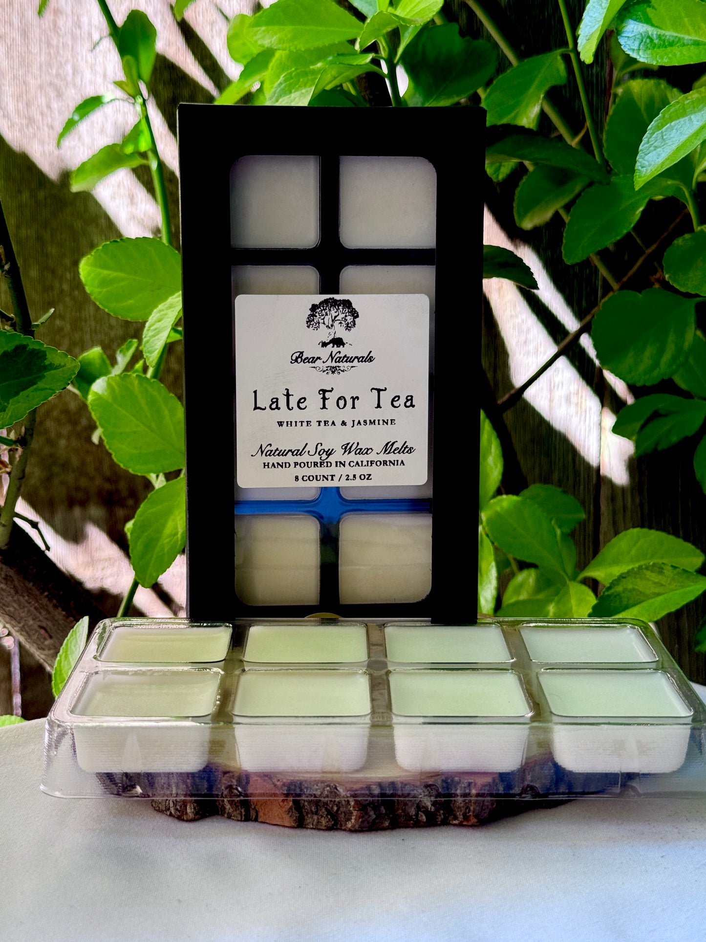 A black box with a clear plastic tray housing eight white wax melts featuring a white label with black writing on a wooden surface with green leaves in the background