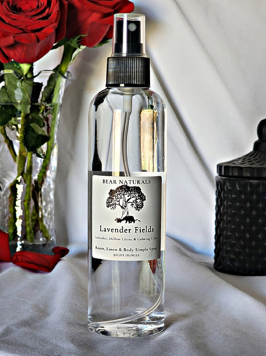 A clear bottle with a black fingertip style spray nozzle featuring a white label with black writing with a black candle jar and red roses in a vase in the background.