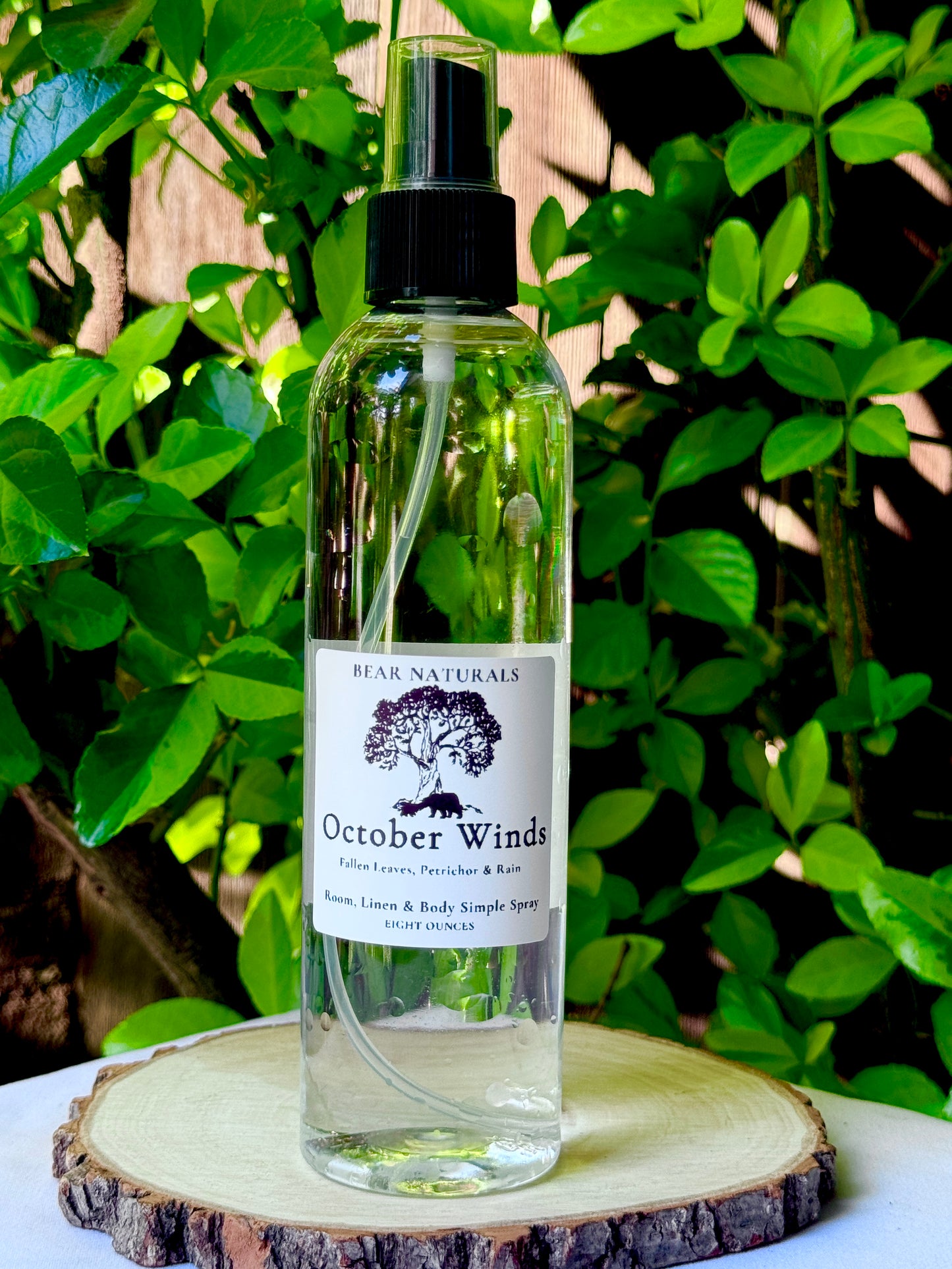 A clear bottle with a black fingertip style spray nozzle featuring a white label with black writing on a wooden surface with green leaves in the background.
