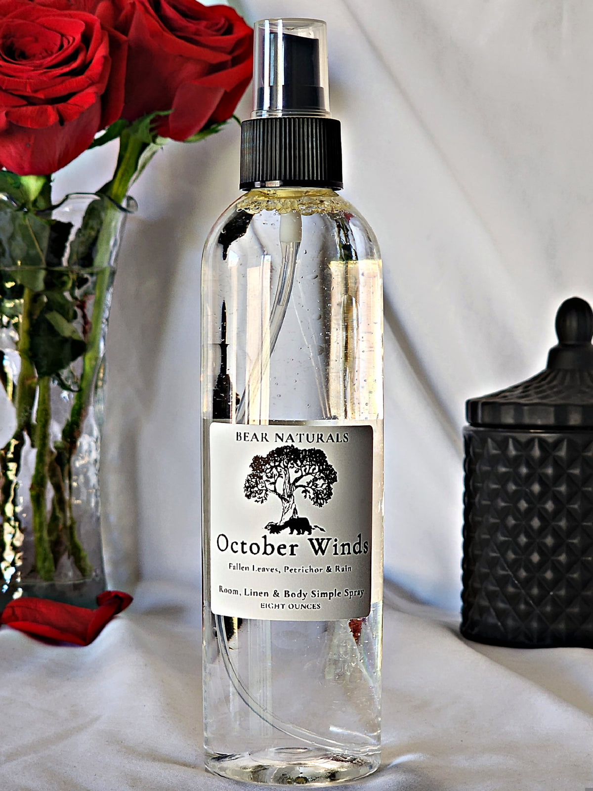 A clear bottle with a black fingertip style spray nozzle featuring a white label with black writing with a black candle jar and red roses in a vase in the background.