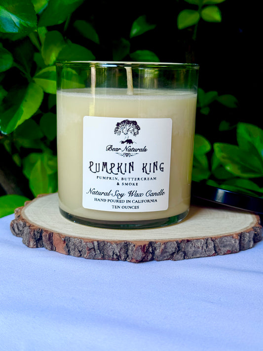 a ten ounce white candle in a clear jar featuring a white label with black writing on a wooden surface with green leaves in the back ground.
