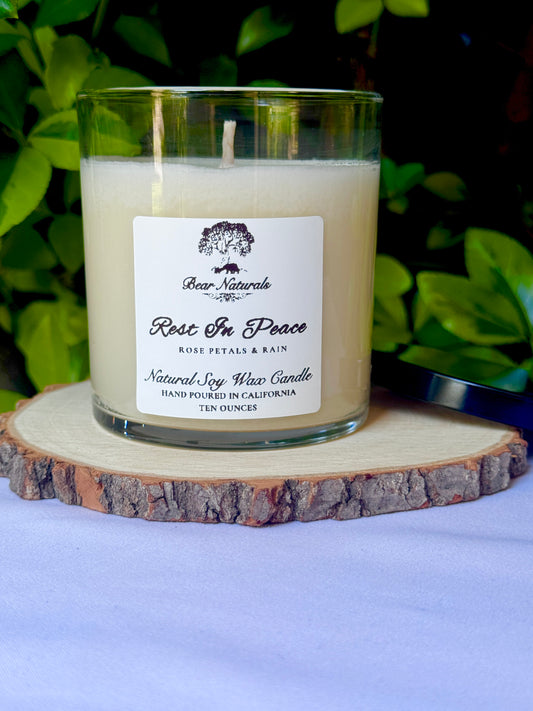 A white ten ounce candle in a clear glass jar with a white label that reads the scent name "Rest In Peace" sitting on a wooden surface with green leaves in the background.