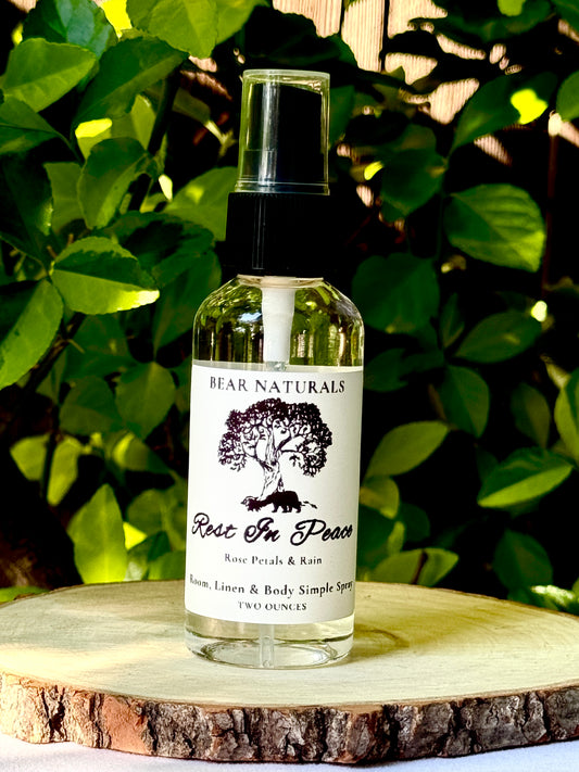 A clear two ounce finger-tip spray bottle with black and white label on a wooden surface with green leaves in the background. The bottle contains the scent "Rest In Peace".