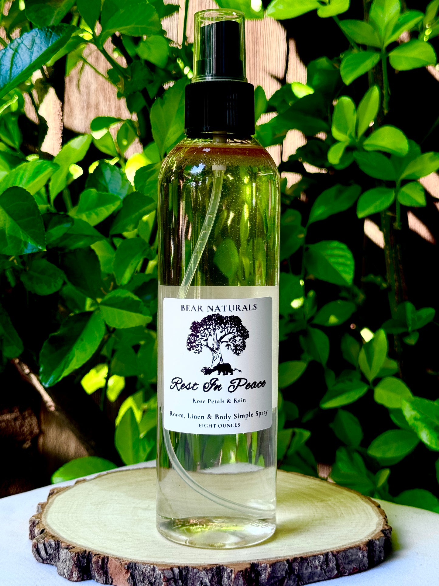 A clear bottle with a black fingertip style spray nozzle featuring a white label with black writing on a wooden surface with green leaves in the background.