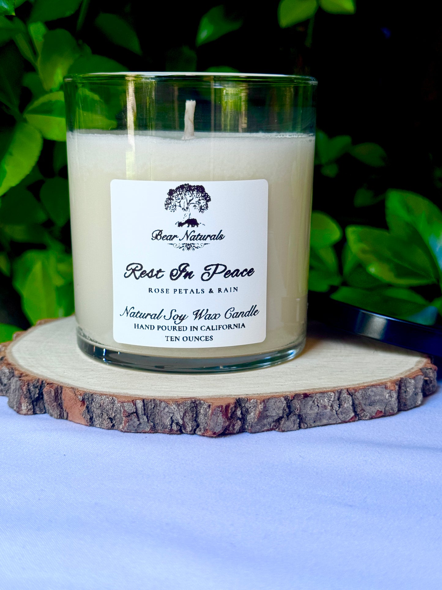 a ten ounce white candle in a clear jar featuring a white label with black writing on a wooden surface with green leaves in the background.