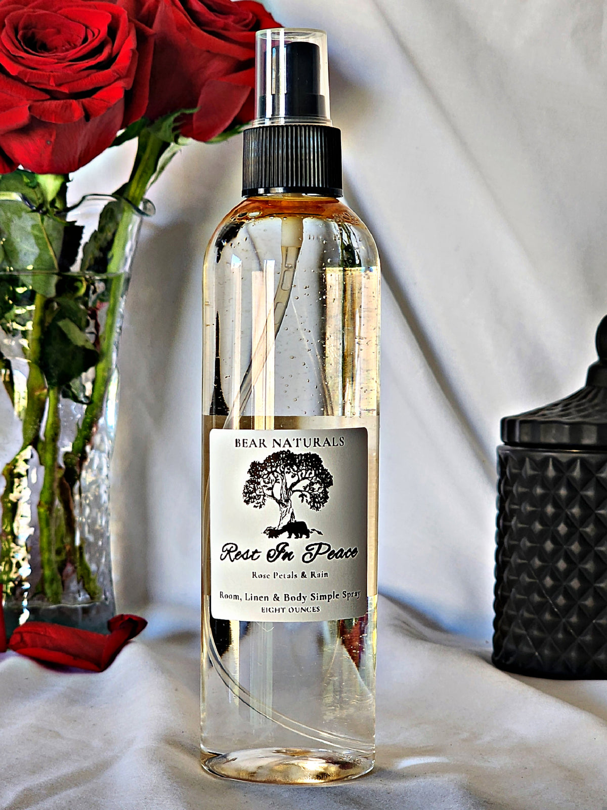 A clear bottle with a black fingertip style spray nozzle featuring a white label with black writing with a black candle jar and red roses in a vase in the background.