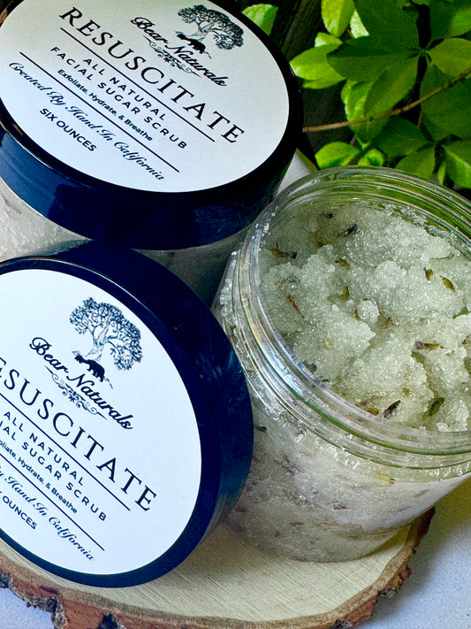 Jar of Resuscitate All Natural Facial scrub with a black lid on a wooden surface with green leaves in the background.