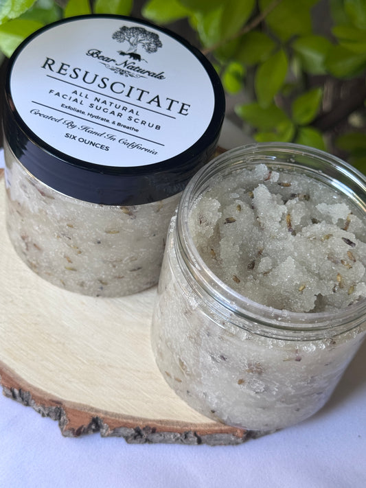 Jar of Resuscitate All Natural Facial scrub with a black lid on a wooden surface with green leaves in the background.