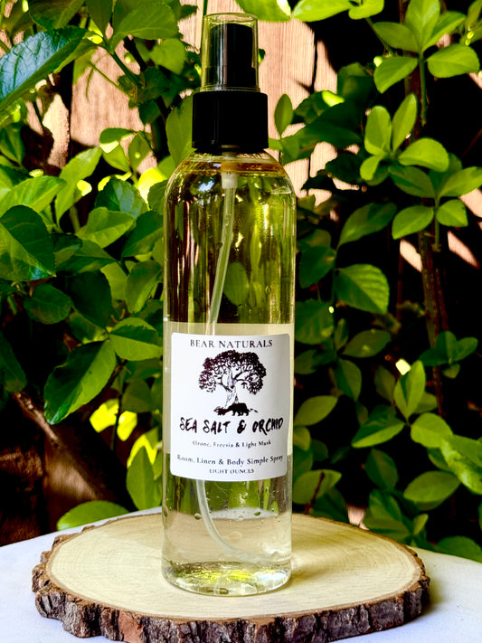 A clear eight ounce finger-tip spray bottle with black and white label on a wooden surface with green leaves in the background. The bottle contains the scent "Sea Salt And Orchid".