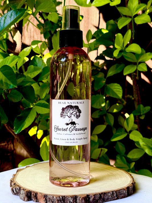 A clear eight ounce finger-tip spray bottle with black and white label on a wooden surface with green leaves in the background. The bottle contains the scent "Secret Passage".
