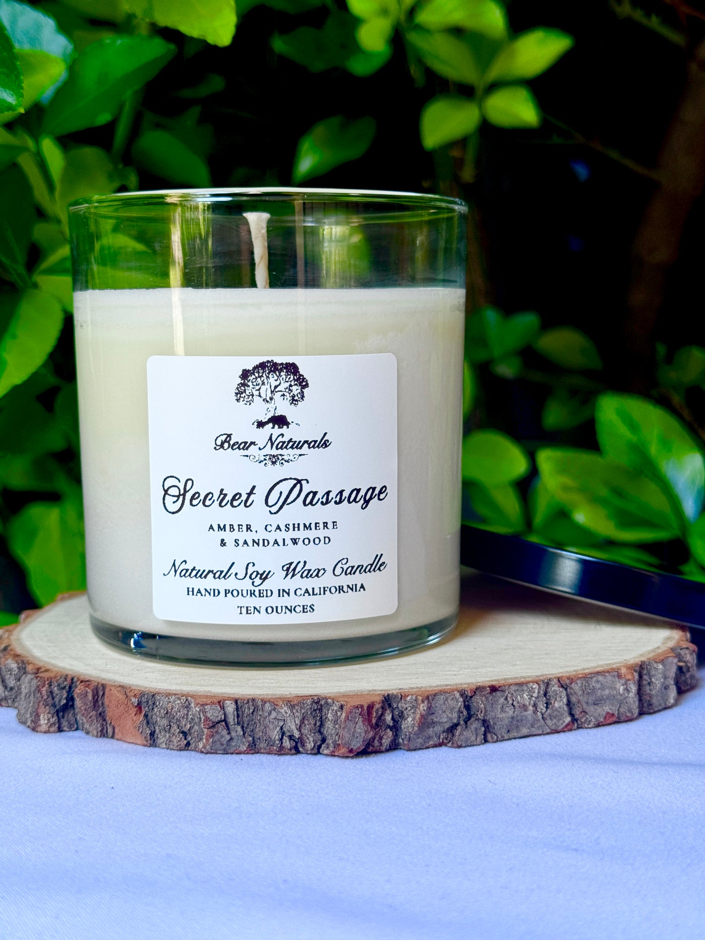a ten ounce white candle in a clear jar featuring a white label with black writing on a wooden surface with green leaves in the back ground.