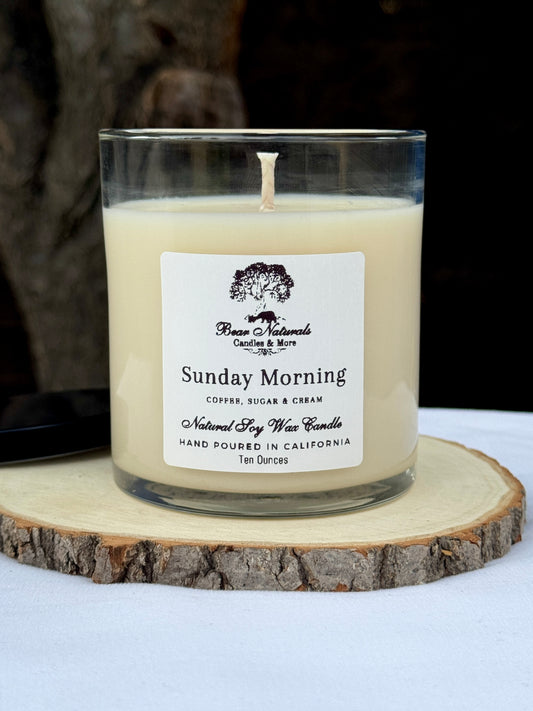 A white ten ounce candle in a clear glass jar with a black metal lid and white label with black writing sitting on a wooden surface. The candle is called "Sunday Morning".