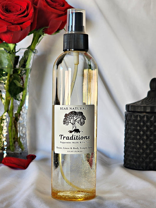 A clear bottle with a black fingertip style spray nozzle featuring a white label with black writing with a black candle jar and red roses in a vase in the background.
