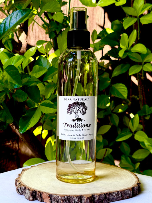 A clear eight ounce finger-tip spray bottle with black and white label on a wooden surface with green leaves in the background. The bottle contains the scent "Traditions".