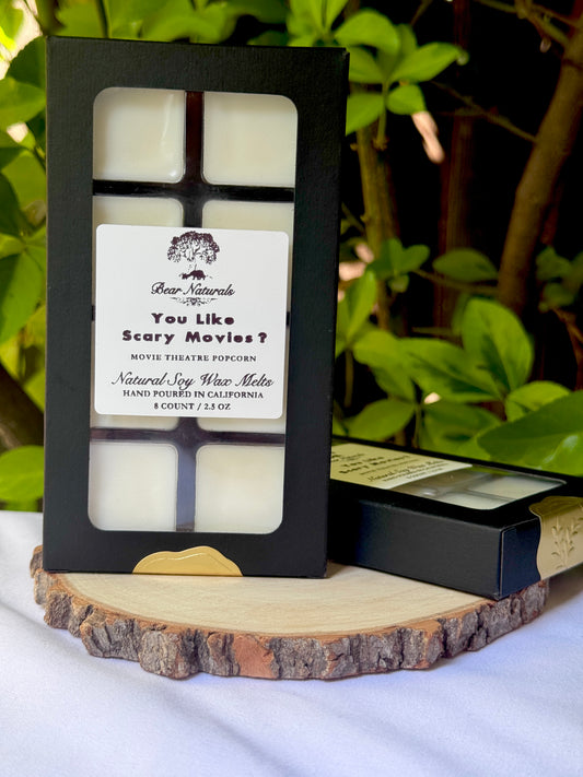 A black box with a clear plastic tray housing eight white wax melts featuring a white label with black writing on a wooden surface with green leaves in the background