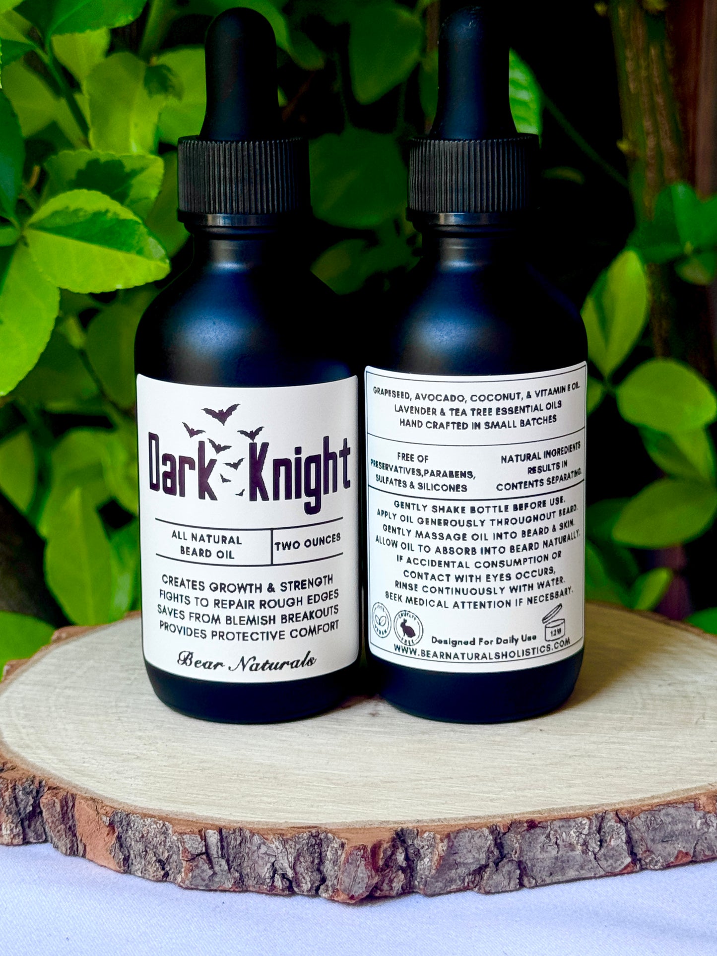 Two, two-ounce black bottles of Dark Knight all-natural beard oil on a wooden surface with green leaves in the background