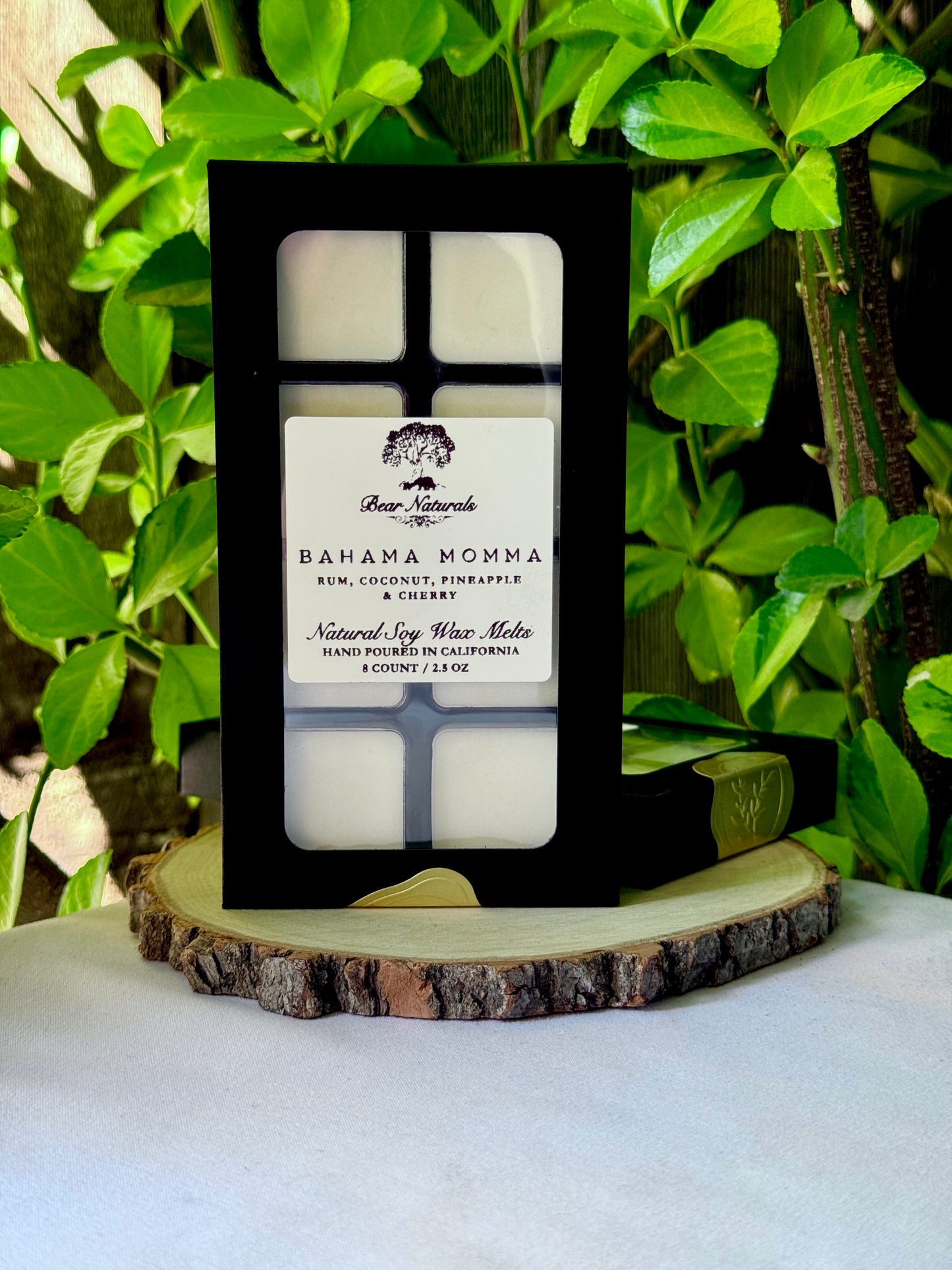 A black box with a clear plastic tray housing eight white wax melts featuring a white label with black writing on a wooden surface with green leaves in the background