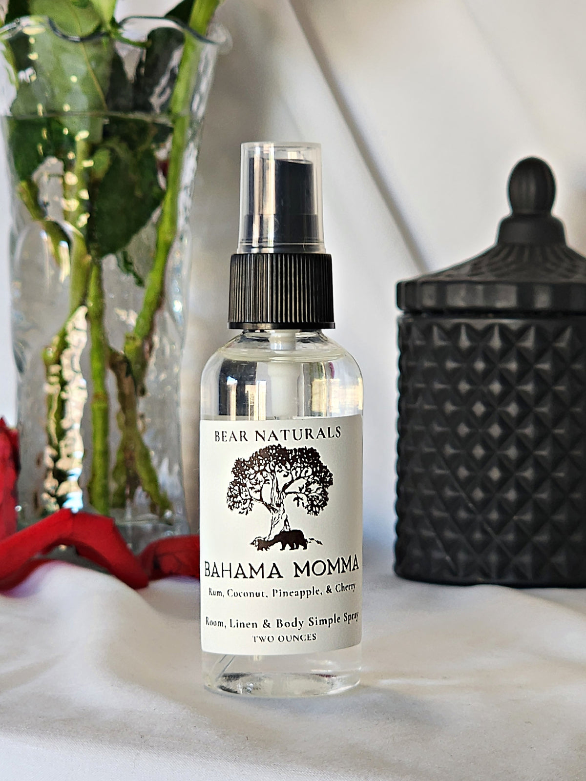 A clear bottle with a black fingertip style spray nozzle featuring a white label with black writing with a black candle jar and red roses in a vase in the background.