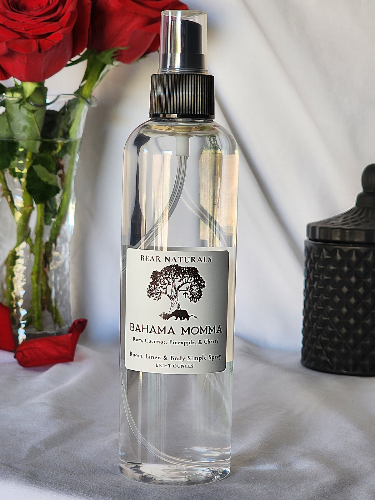 A clear bottle with a black fingertip style spray nozzle featuring a white label with black writing with a black candle jar and red roses in a vase in the background.