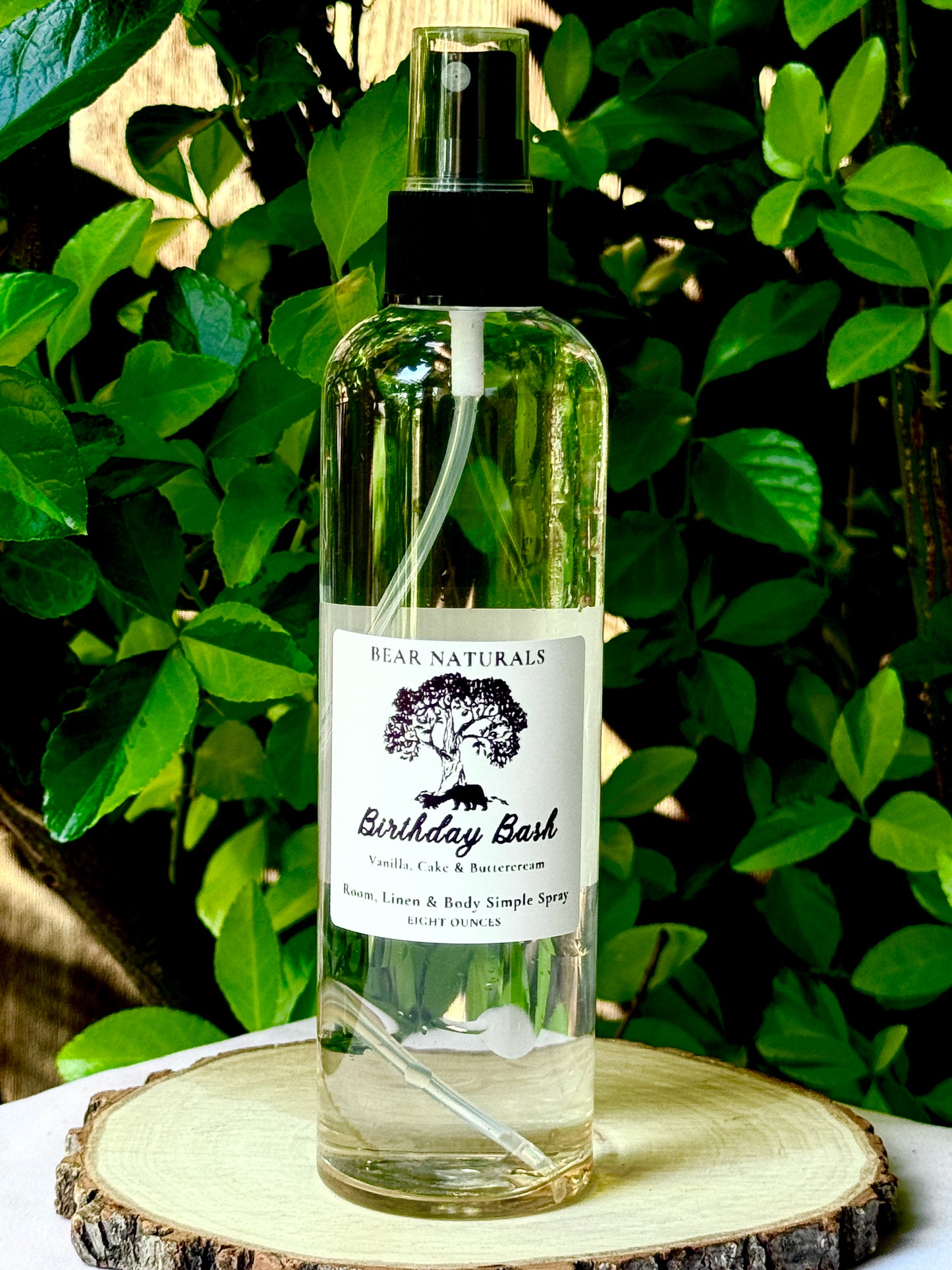 A clear bottle with a black fingertip style spray nozzle featuring a white label with black writing on a wooden surface with green leaves in the background.
