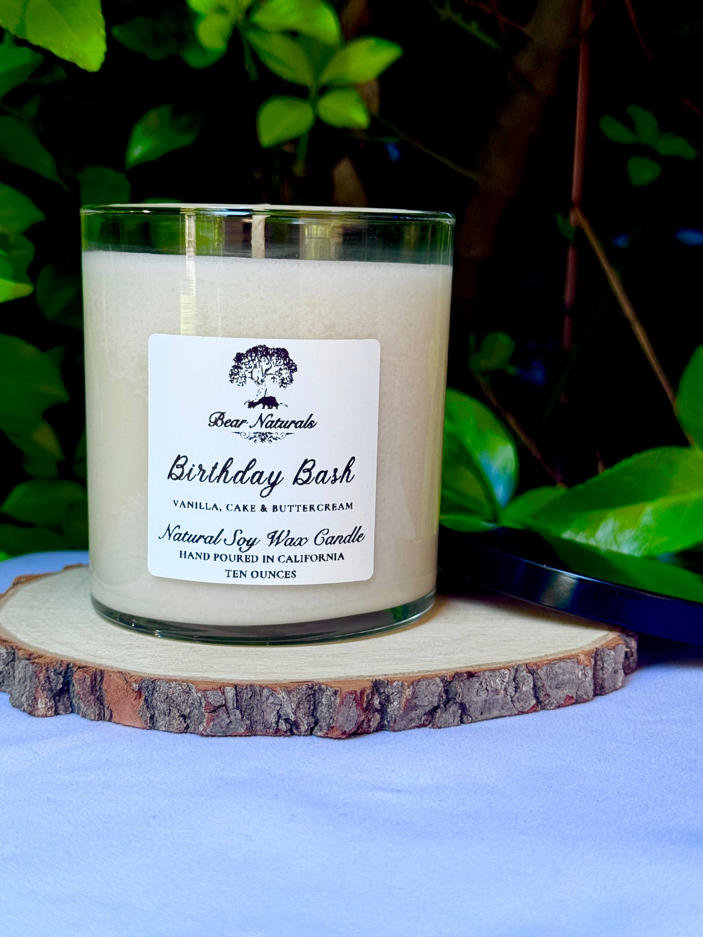 a ten ounce white candle in a clear jar featuring a white label with black writing on a wooden surface with green leaves in the back ground.