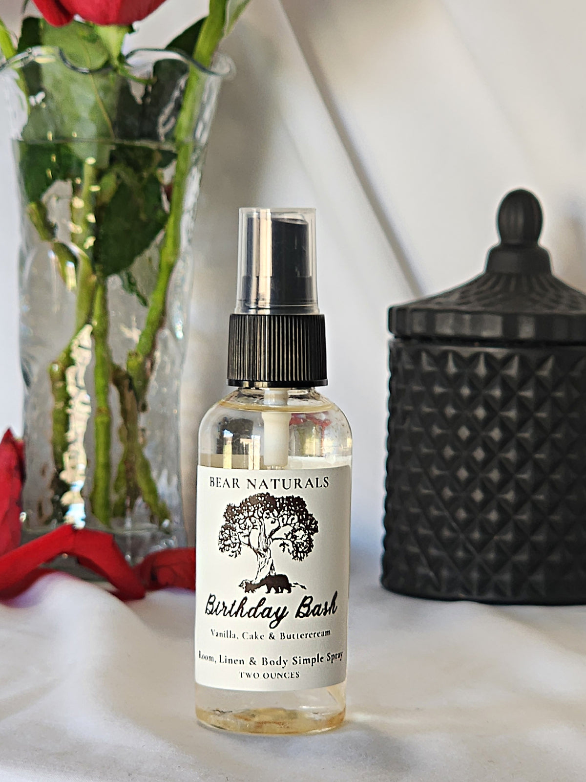 A clear bottle with a black fingertip style spray nozzle featuring a white label with black writing with a black candle jar and red roses in a vase in the background.