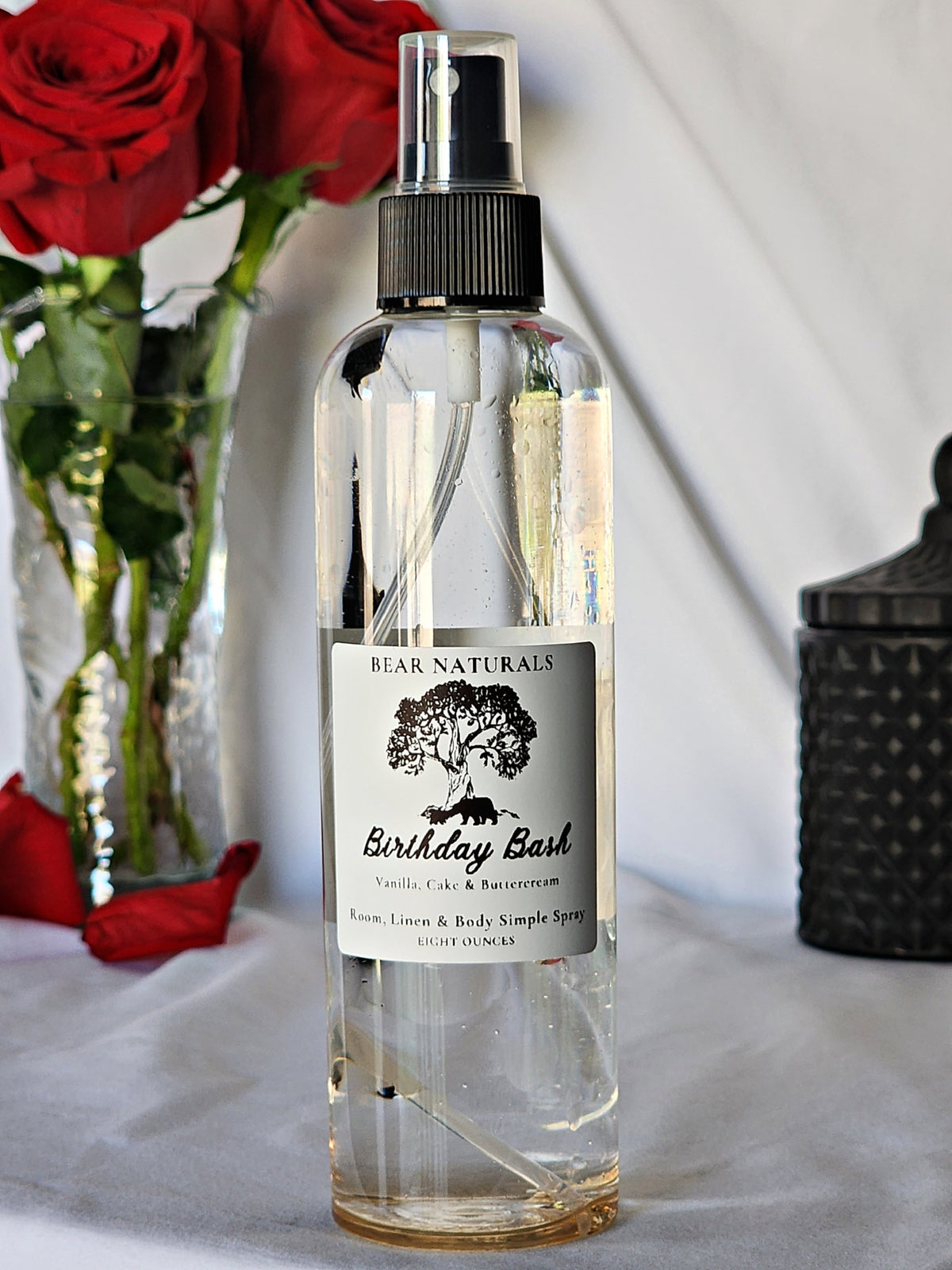 A clear bottle with a black fingertip style spray nozzle featuring a white label with black writing on a wooden surface with green leaves in the background.