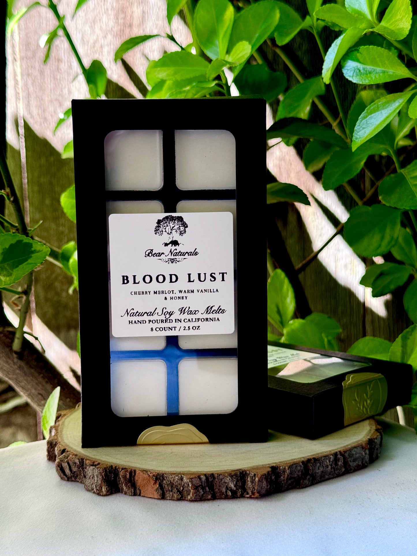 A black box with a clear plastic tray housing eight white wax melts featuring a white label with black writing on a wooden surface with green leaves in the background