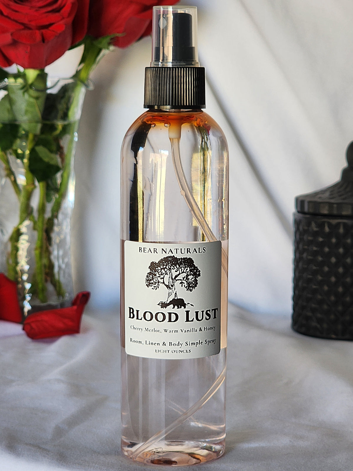 A clear bottle with a black fingertip style spray nozzle featuring a white label with black writing with a black candle jar and red roses in a vase in the background.