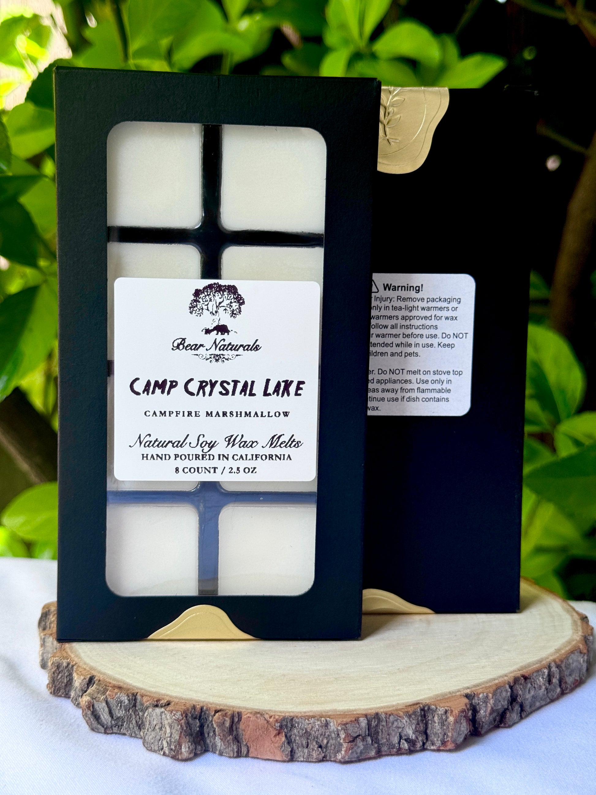 A black box with a clear plastic tray housing eight white wax melts featuring a white label with black writing on a wooden surface with green leaves in the background