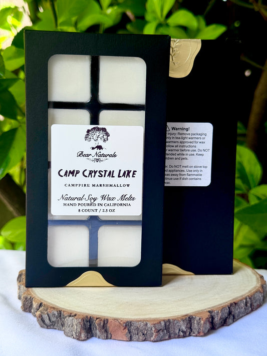 A black box with a clear plastic tray housing eight white wax melts featuring a white label with black writing on a wooden surface with green leaves in the background