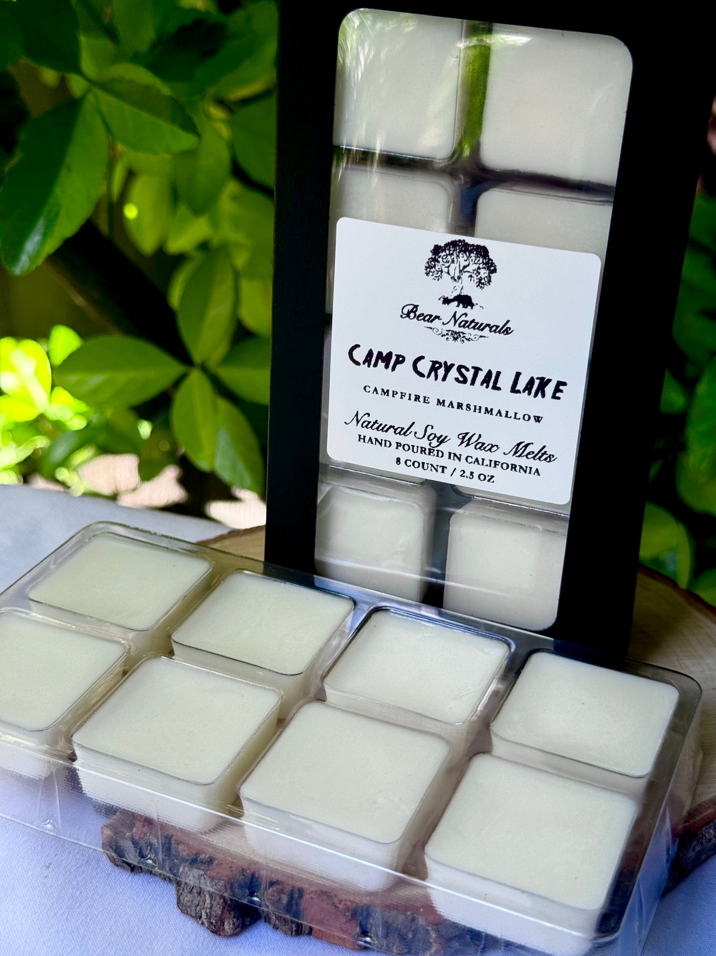 A black box with a clear plastic tray housing eight white wax melts featuring a white label with black writing on a wooden surface with green leaves in the background