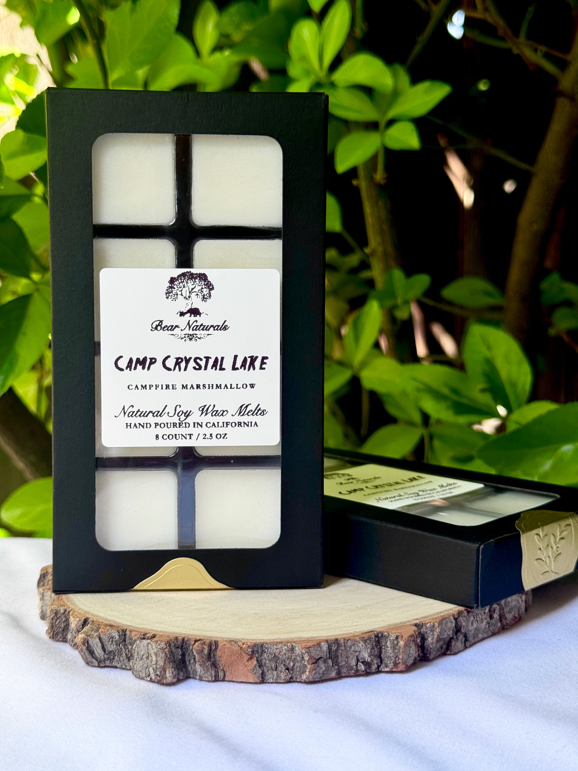A black box with a clear plastic tray housing eight white wax melts featuring a white label with black writing on a wooden surface with green leaves in the background