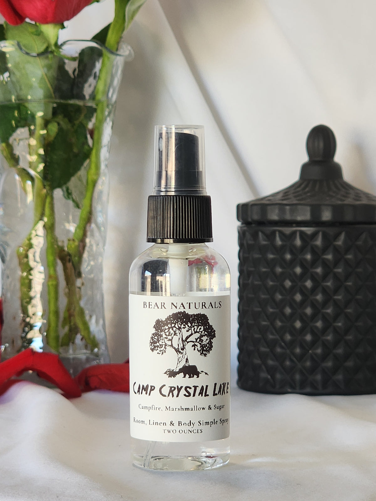 A clear bottle with a black fingertip style spray nozzle featuring a white label with black writing with a black candle jar and red roses in a vase in the background.