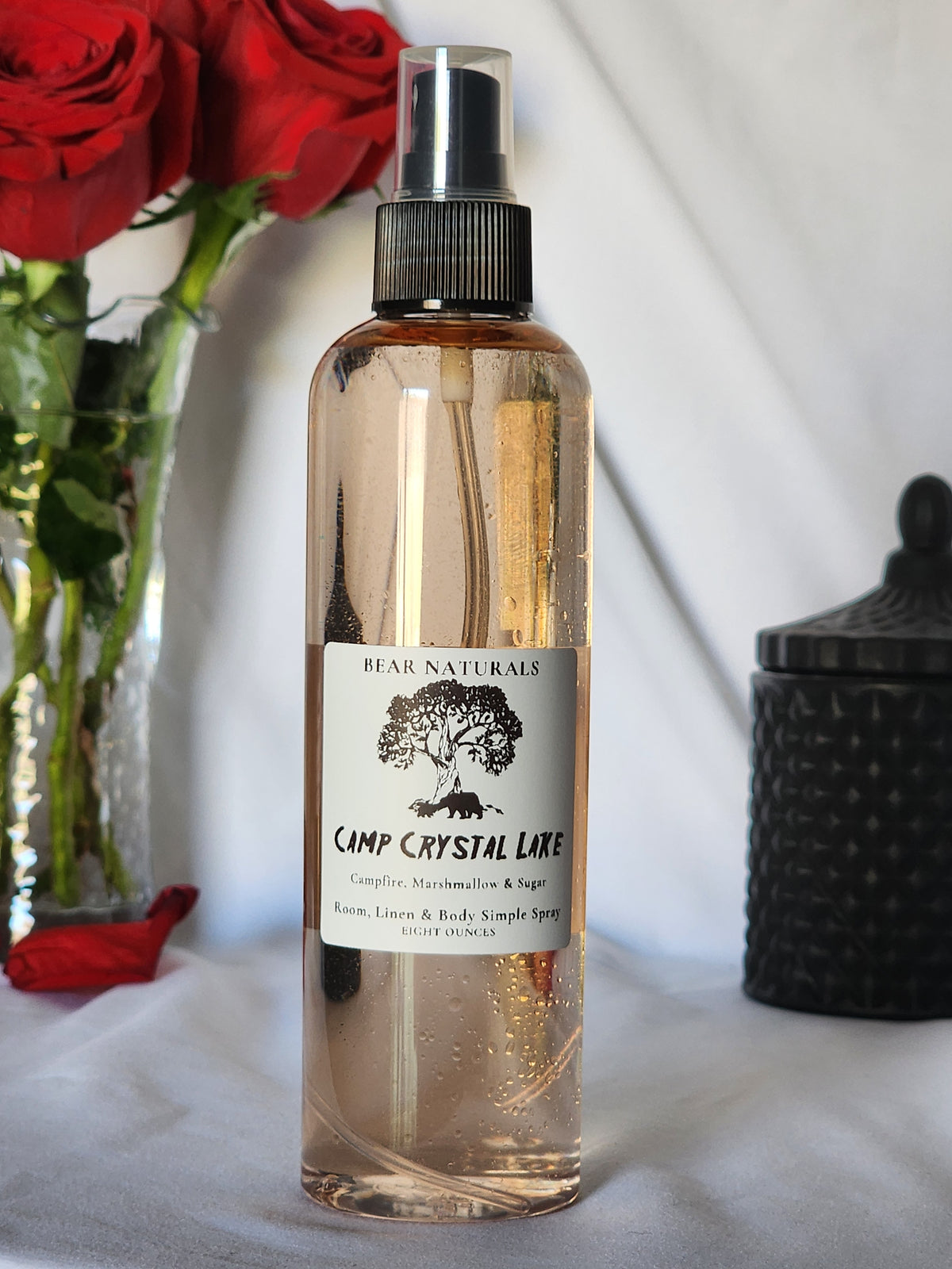 A clear bottle with a black fingertip style spray nozzle featuring a white label with black writing with a black candle jar and red roses in a vase in the background.