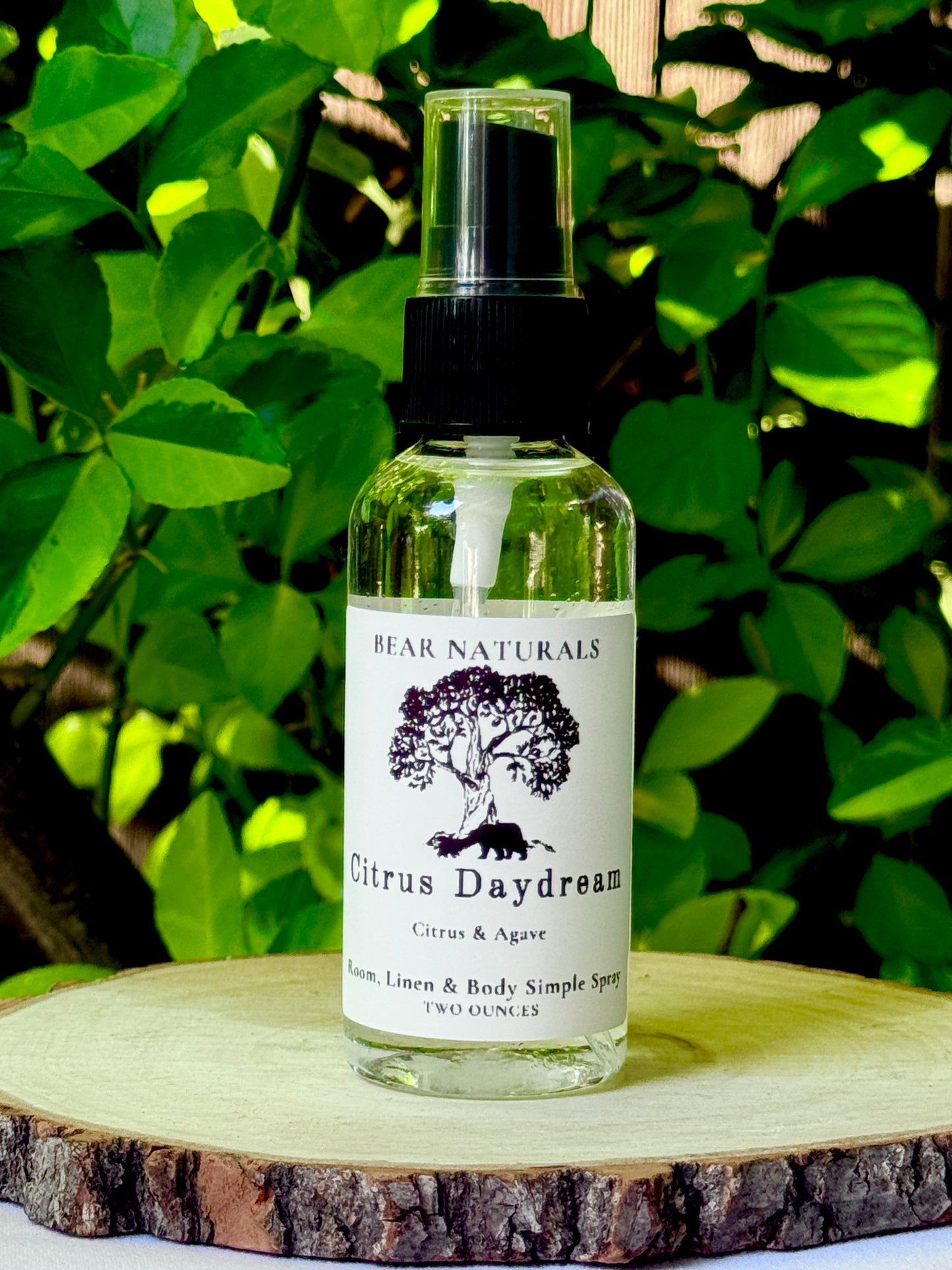 A clear bottle with a black fingertip style spray nozzle featuring a white label with black writing on a wooden surface with green leaves in the background.