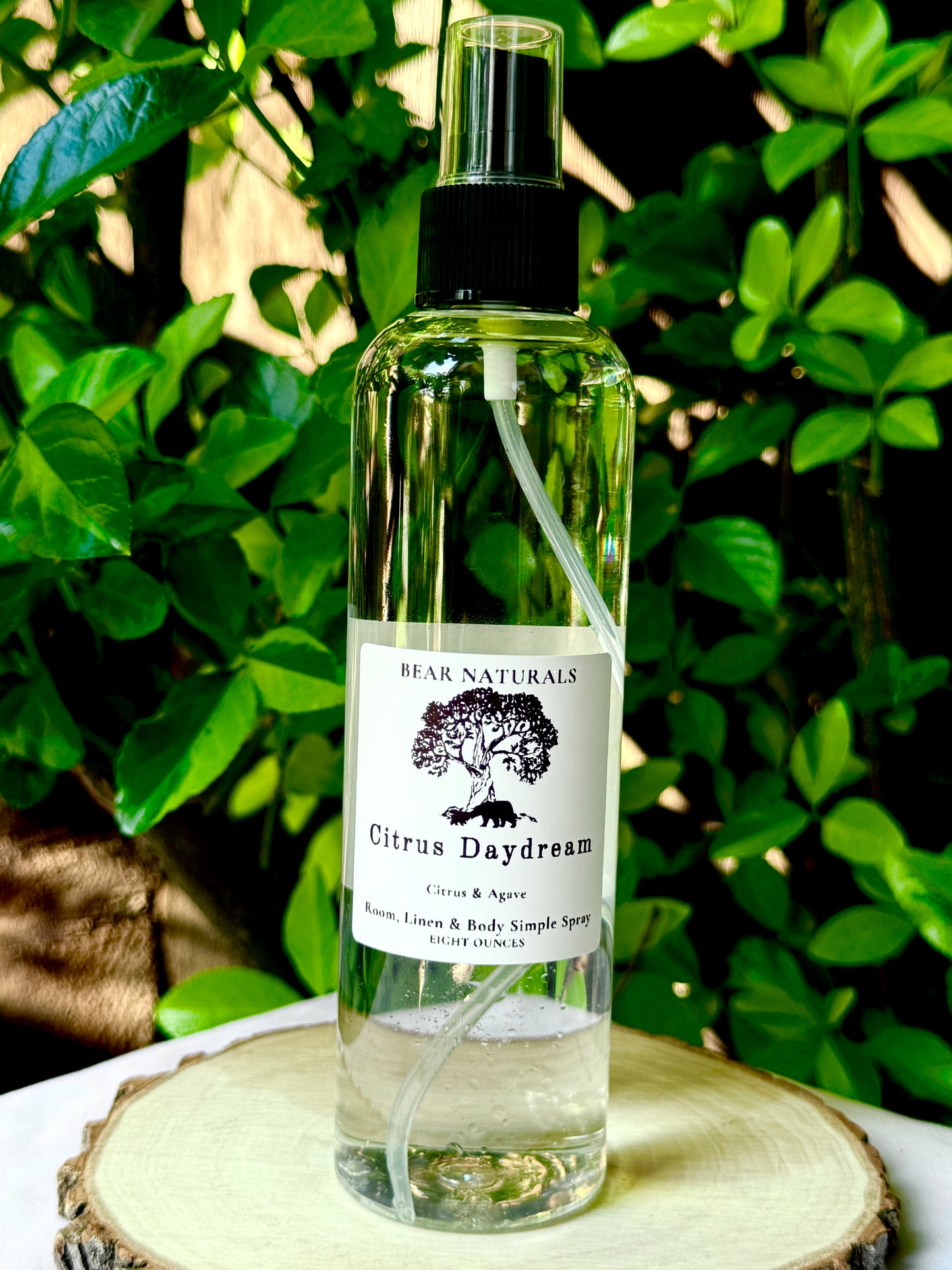 A clear bottle with a black fingertip style spray nozzle featuring a white label with black writing on a wooden surface with green leaves in the background.