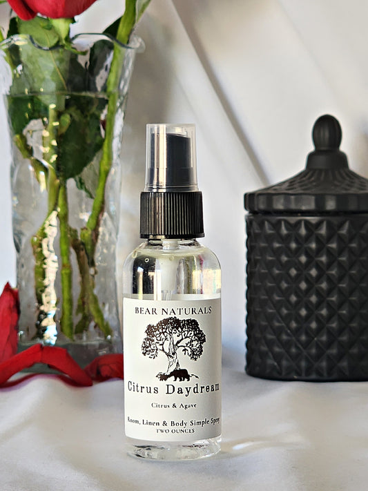 A clear bottle with a black fingertip style spray nozzle featuring a white label with black writing with a black candle jar and red roses in a vase in the background.