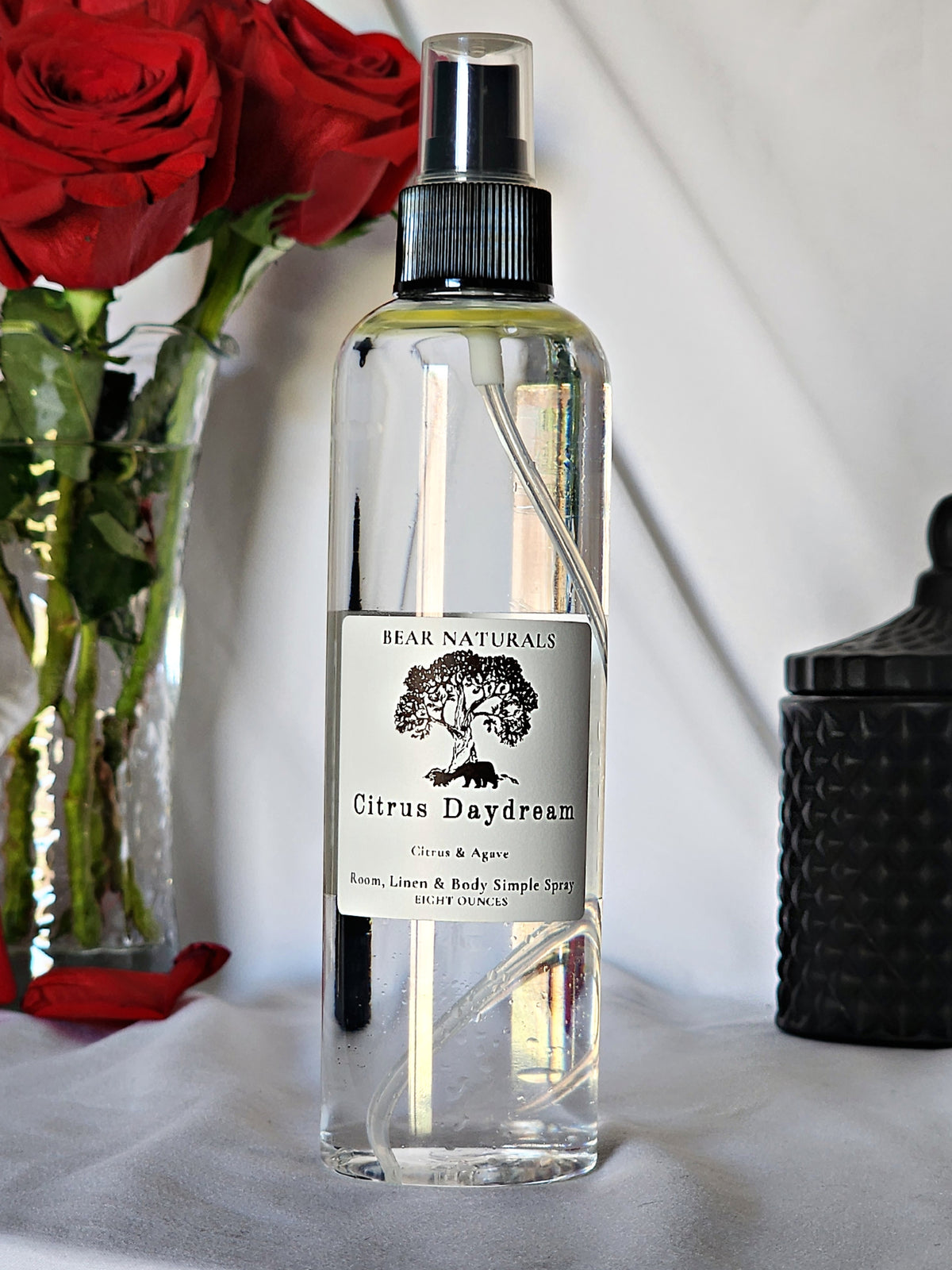 A clear bottle with a black fingertip style spray nozzle featuring a white label with black writing with a black candle jar and red roses in a vase in the background.